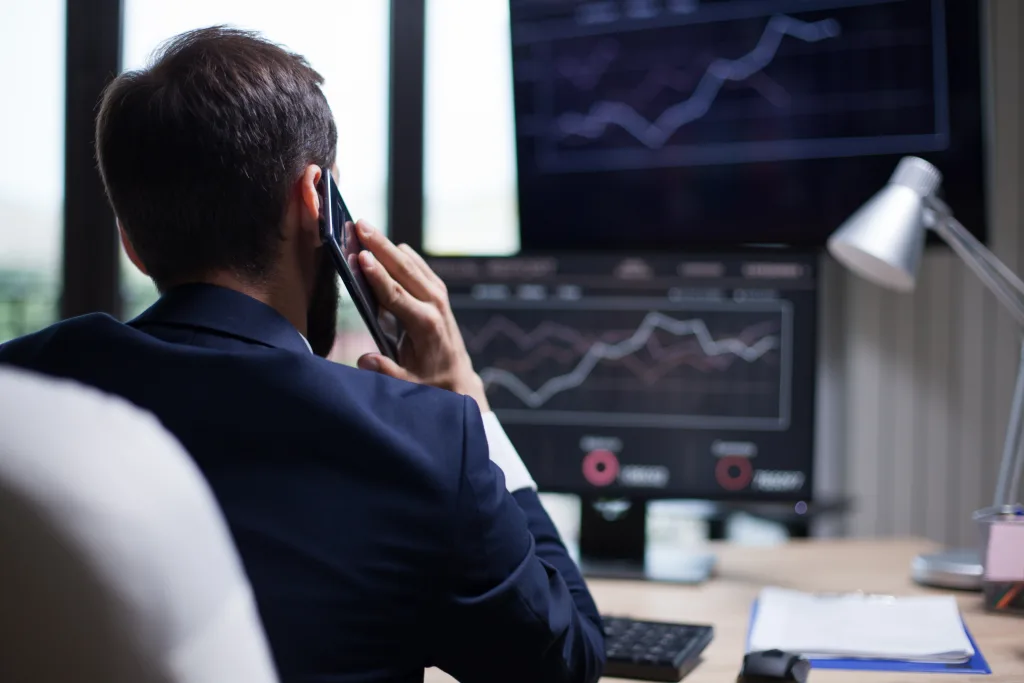 US financial analyst on a phone call while monitoring stock market trends and charts on dual screens in a modern office setting.