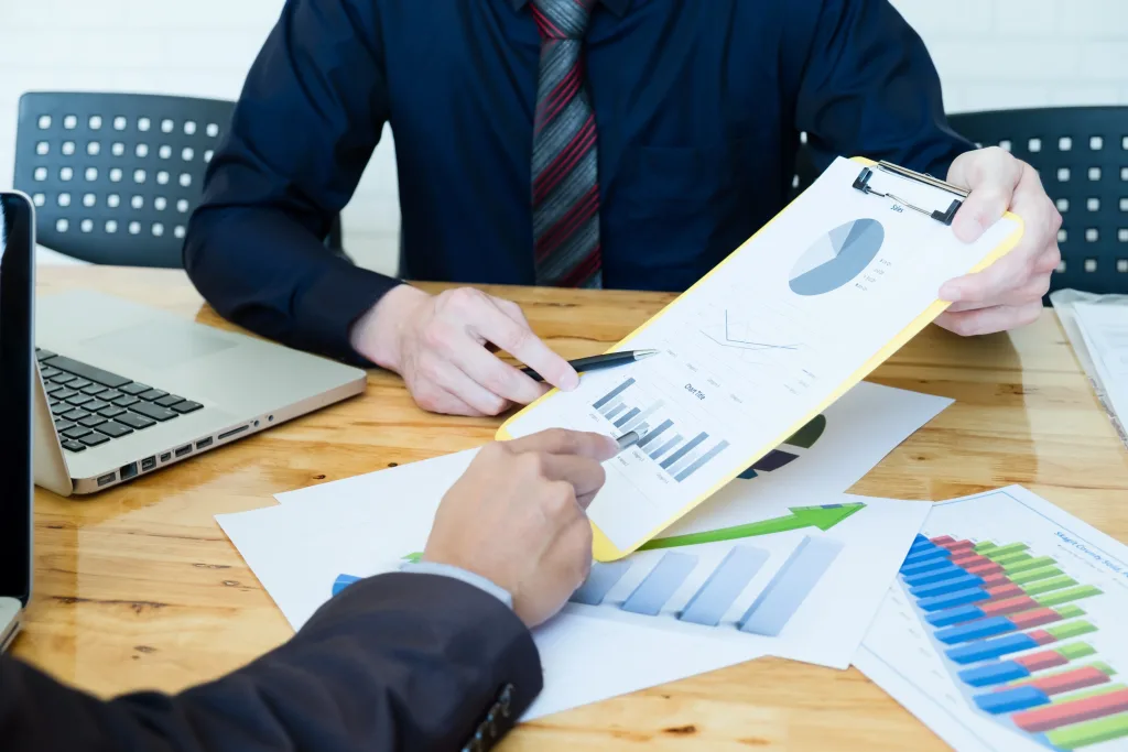 A close-up shot of two individuals at a wooden table, engaged in a business discussion involving documents and a laptop. One person, wearing a dark shirt and patterned tie, holds a yellow clipboard with a chart featuring a pie graph and bar charts, pointing to it with a pen. The other person, wearing a dark suit jacket, points to a separate document on the table that displays a green upward-sloping arrow graph and a multi-colored bar chart. This scene suggests a meeting focused on reviewing financial data, likely for annual ASIC compliance management, including preparing statements, filing declarations, and updating records.