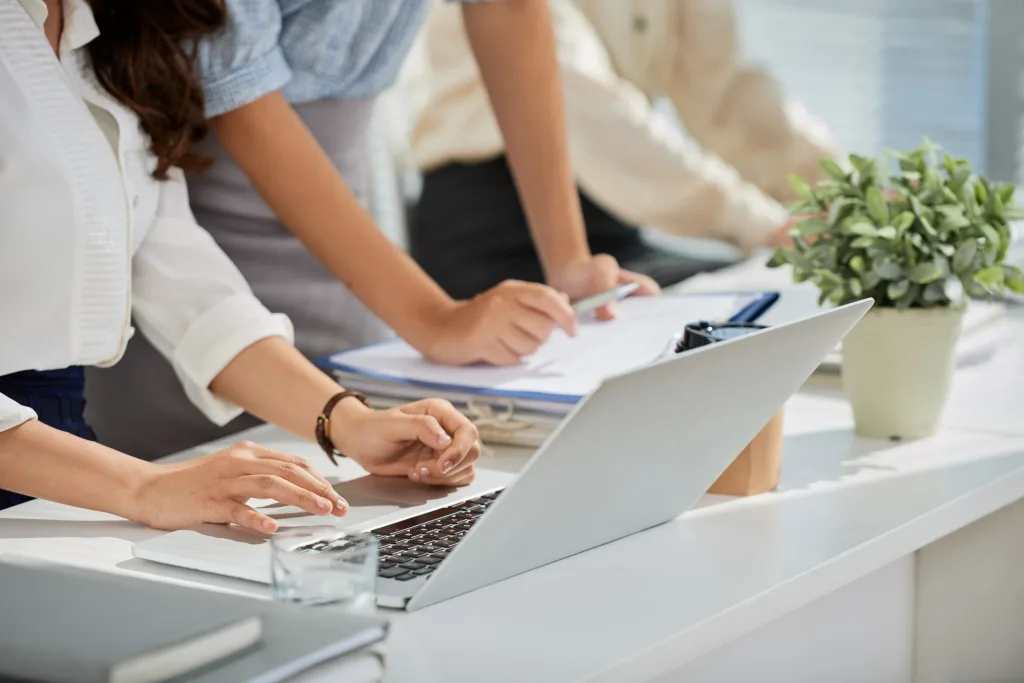A dynamic shot captures two individuals, likely colleagues, collaborating at a white desk in an office setting. In the foreground, a person is seated, with their hands positioned on a silver laptop keyboard, actively working. Beside the laptop is a glass of water and some stacked books. Another person stands slightly behind and to the right, leaning over and pointing with a pen at documents on a clipboard, suggesting a discussion or review of paperwork. The background shows a third person faintly, also appearing to be engaged in work, and a potted plant adds a touch of greenery to the professional environment. This scene depicts teamwork and administrative tasks, possibly related to business name registration or maintaining a registered office, as implied by the accompanying text.