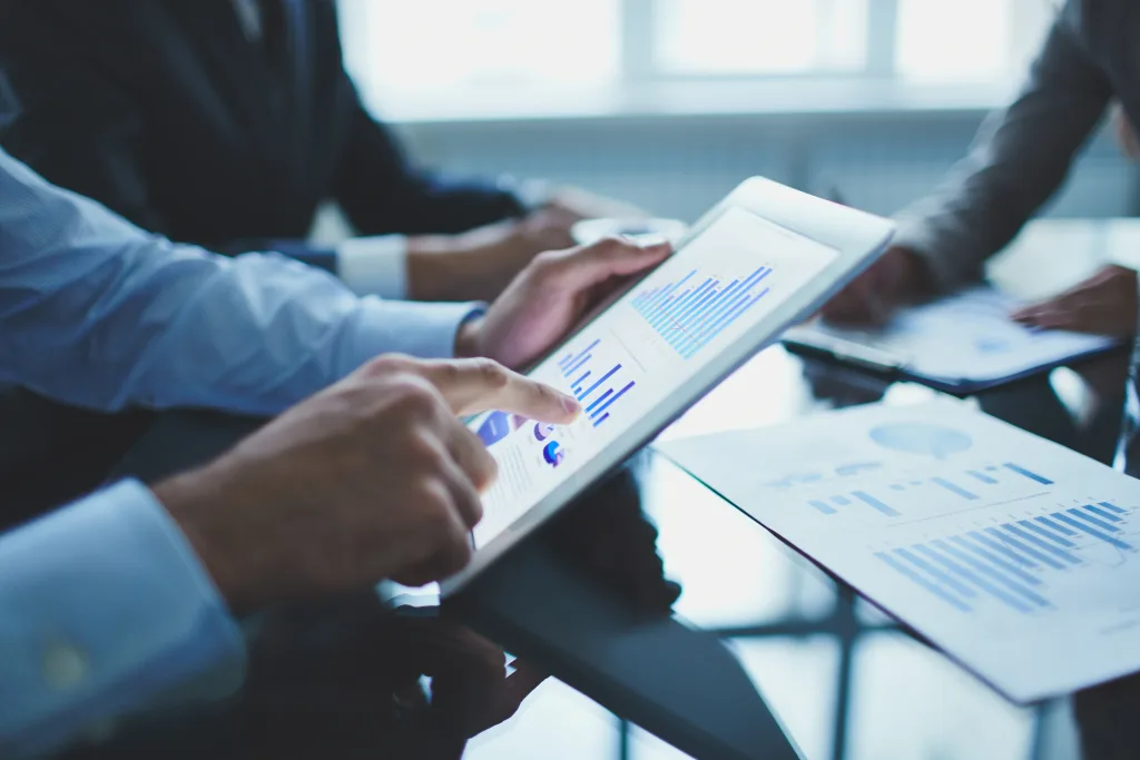 A close-up shot captures a person in a light blue shirt actively engaging with a white tablet, displaying various blue bar graphs and charts. Their finger is poised over the screen, indicating interaction with the data. On the dark table, a paper document with similar blue graphs is also visible. In the blurred background, other individuals in business attire suggest a collaborative meeting or discussion. The scene emphasizes data analysis and digital tools being used for compliance monitoring and strategic planning, likely to help avoid late fees, delays, and legal risks, as the accompanying text implies.