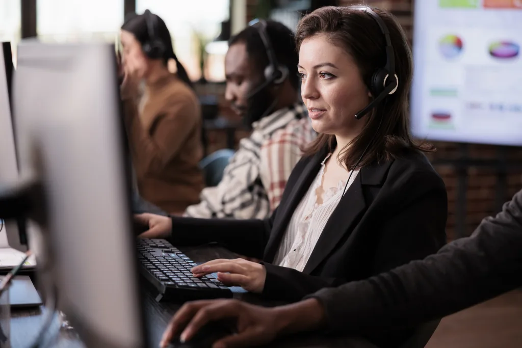 A diverse group of customer service representatives, each wearing a headset, are seated at individual computer workstations in an office setting. In the foreground, a woman with dark hair in a black blazer and white patterned blouse is intently focused on her computer screen, with her hands on the keyboard. Behind her, other colleagues, including a man and another person in the background, are also working at their stations, wearing headsets. A large screen displaying charts and graphs is visible in the background, suggesting a data-driven or call center environment. The scene depicts a busy and professional call center or corporate secretarial support service, where agents are likely handling inquiries and administrative tasks, such as those related to ASIC agent services, drafting corporate documents, or maintaining statutory records, as described in the accompanying text.